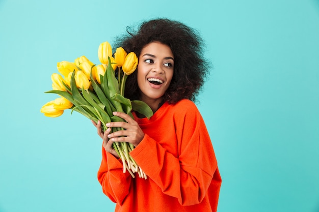 Gelockte amerikanische Frau in der roten Kleidung, die beiseite schaut und Bündel der schönen gelben Tulpen hält, lokalisiert über blaue Wand