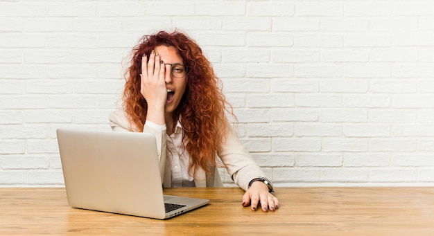 Gelockte Frau der jungen Rothaarigen, die mit ihrem Laptop hat den Spaß bedeckt Hälfte des Gesichtes mit Palme arbeitet.