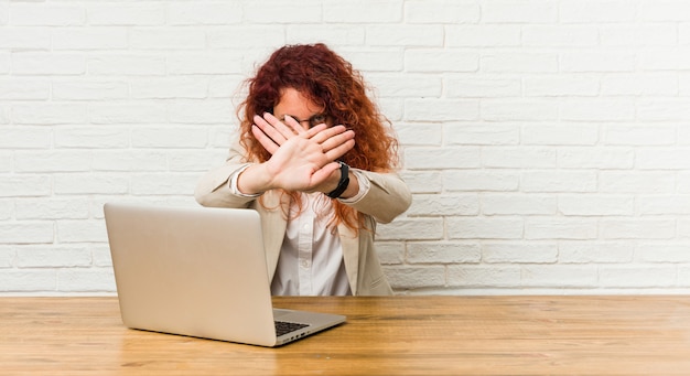 Gelockte Frau der jungen Rothaarigen, die mit ihrem Laptop tut eine Ablehnungsgeste arbeitet