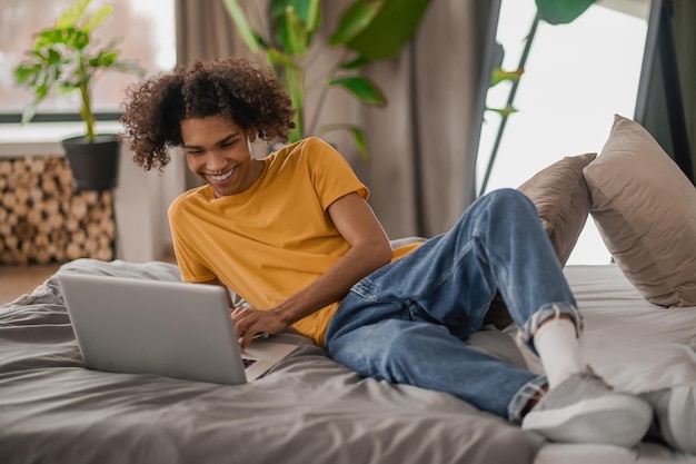 Gelockter junger Kerl, der auf einem Bett mit einem Laptop sitzt