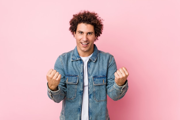 Gelockter reifer Mann, der eine Denimjacke gegen das rosa Hintergrundzujubeln sorglos und aufgeregt trägt. Sieg-Konzept.