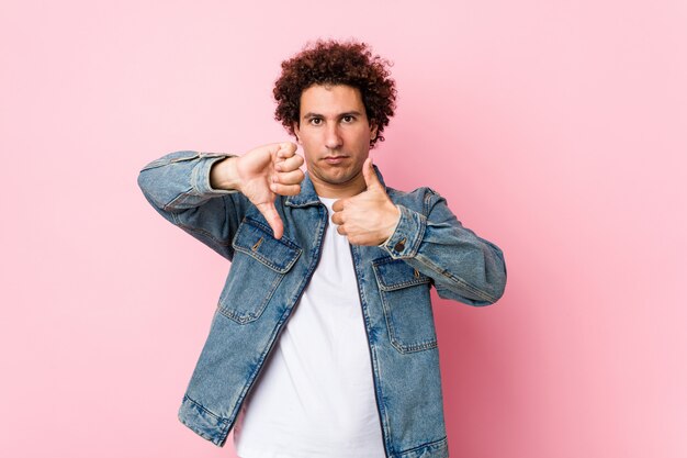 Gelockter reifer Mann, der eine Denimjacke gegen den rosa Hintergrund sich zeigt Daumen und Daumen unten, wählen schwieriges Konzept trägt