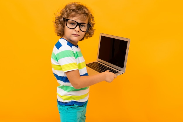 Gelockter rothaariger Junge mit Gläsern in einem gestreiften T-Shirt, das einen Laptop in seinen Händen auf einem gelben Hintergrund hält