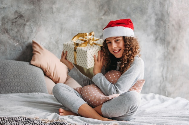 Gelocktes behaartes schönes Tweenmädchen in Sankt-Hut und in Pyjamas, die auf Bett mit großer Geschenkbox, Weihnachtsmorgenzeit sitzen