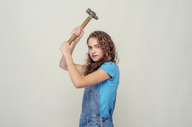 Gelocktes, fröhliches, brünettes Mädchen in Denim-Overalls hält Hammer in der Hand und hebt ihn über Kopfschaukeln