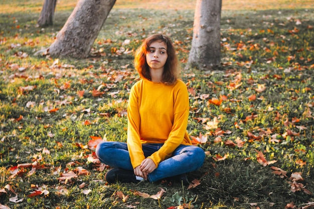 Gelocktes junges Mädchen in gelbem Pullover und Jeans, das auf Herbstgras mit trockenen Blättern sitzt. Herbststimmung