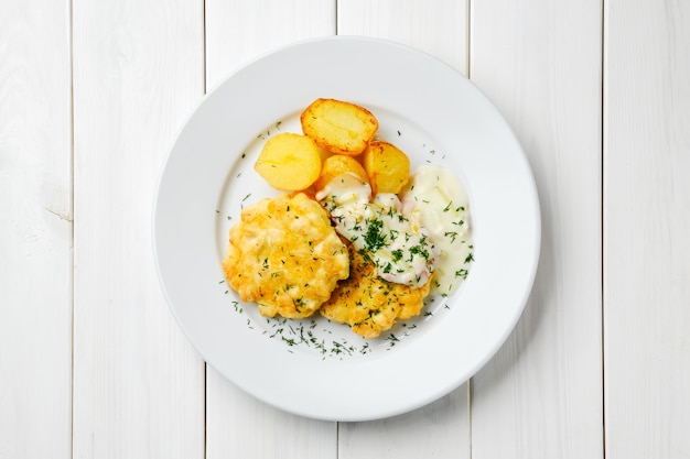 Gemahlenes Hühnerfleischkotelett mit gebratenen Kartoffelschnitzen und cremiger Sauce mit Speck auf weißem Holztisch