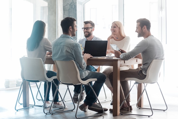 Gemeinsam an einem neuen Projekt arbeiten. Gruppe selbstbewusster Geschäftsleute in smarter Freizeitkleidung, die zusammenarbeiten, während sie im Büro am Schreibtisch sitzen