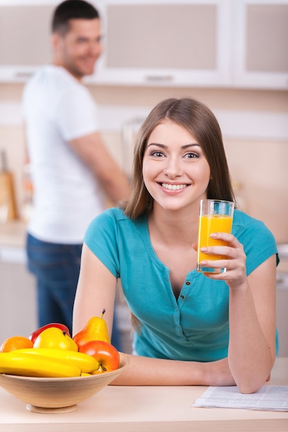 Gemeinsam den Sonntagmorgen verbringen. Schöne junge Frau, die sich an den Küchenherd lehnt und ein Glas mit Orangensaft hält, während der Mann im Hintergrund steht und lächelt