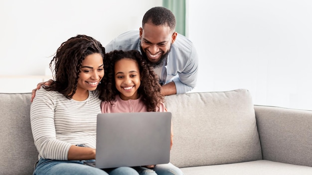 Gemeinsam entspannen. Portrait von glücklichen schwarzen Eltern und ihrer süßen Tochter, die Laptop benutzen, Zeit miteinander verbringen, Filme schauen oder im Internet surfen, auf dem Sofa im Wohnzimmer sitzen, Platz kopieren, Banner