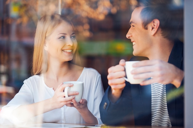 Gemeinsam frischen Kaffee genießen. Durch eine Glasaufnahme eines schönen jungen Paares, das sich anschaut und lächelt, während es zusammen Kaffee im Café genießt?