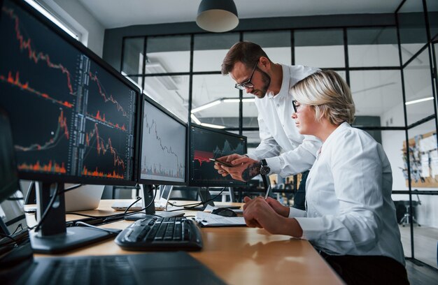 Gemeinsam Informationen analysieren. Zwei Börsenmakler in formeller Kleidung arbeiten im Büro mit Finanzmarkt.