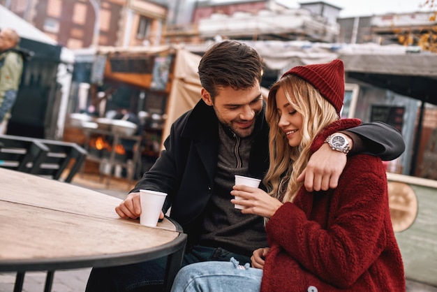 Gemeinsam Kaffee trinken. Junge Paare sprechen beim Trinken von heißem Getränk, Tee oder Kaffee im Freien. Sie sitzen auf der Bank, junger Mann umarmt seine Freundin