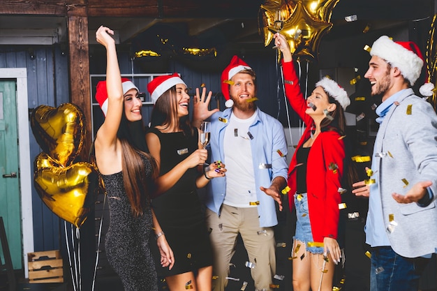 Gemeinsam Neujahr feiern. Gruppe der schönen jungen Leute in den Weihnachtsmützen, die bunte Konfetti werfen und glücklich schauen