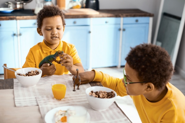 Gemeinsam Spaß haben. Charmante kleine Jungen, die mit ihren Spielzeugdinosauriern spielen, während sie am Tisch sitzen und Müsli essen