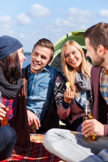 Gemeinsam tolle Zeit verbringen. Gruppe junger fröhlicher Leute, die beim Campen am Flussufer Bier trinken und reden?