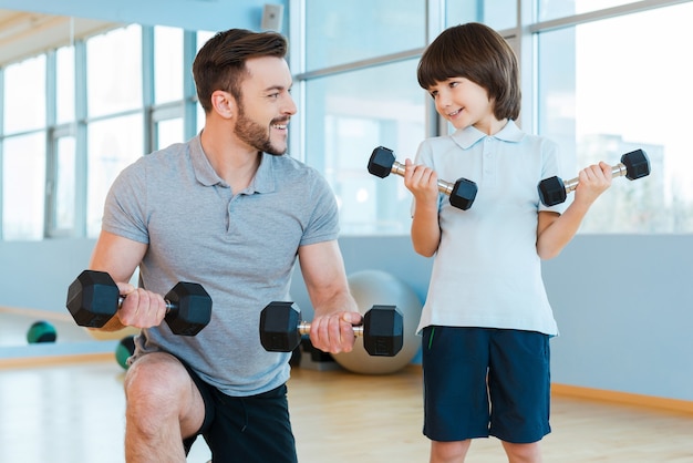 Gemeinsam trainieren. Glücklicher Vater und Sohn, die mit Hanteln trainieren und lächeln, während sie im Fitnessstudio stehen