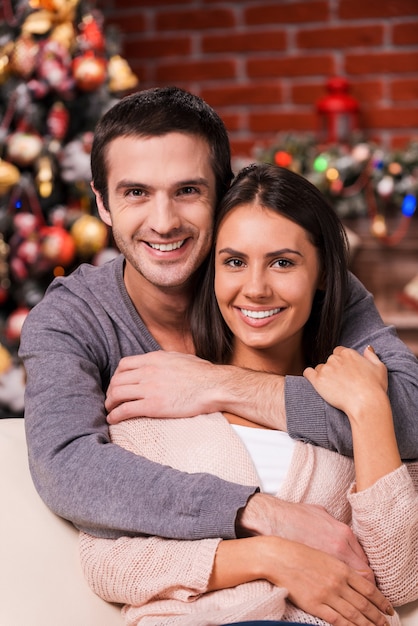 Gemeinsam Weihnachten genießen. Schönes junges Liebespaar, das sich aneinander bindet und mit Weihnachtsbaum im Hintergrund lächelt