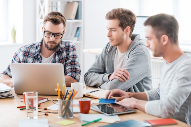 Gemeinsam werden sie es herausfinden. Drei junge selbstbewusste Männer arbeiten zusammen, während sie an ihrem Arbeitsplatz im Büro sitzen