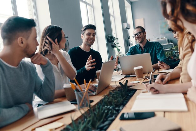 Gemeinsam zum Erfolg. Gruppe junger moderner Menschen in eleganter Freizeitkleidung, die etwas diskutieren und lächeln, während sie im Kreativbüro arbeiten working