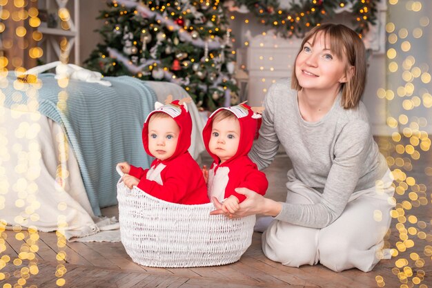 Los gemelos del niño en rojo reno trajes de santa claus están sentados en casa con su madre en el contexto del árbol de Navidad