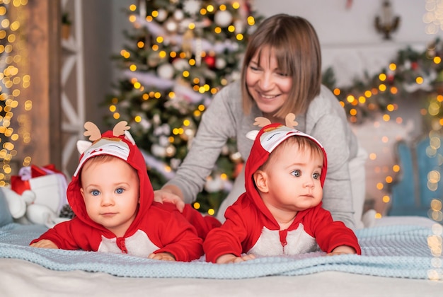Gêmeos bebês com fantasias de Papai Noel de rena vermelha estão sentados em casa com a mãe no fundo da árvore de Natal