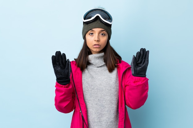 Gemischte Rennskifahrerin mit Snowboardbrille über blauer Wand macht Stoppgeste und enttäuscht