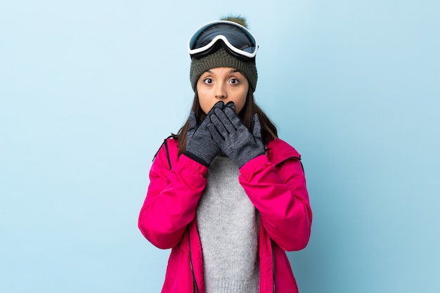 Gemischte Rennskifahrerin mit Snowboardbrille über der blauen Wand, die Mund mit Händen bedeckt