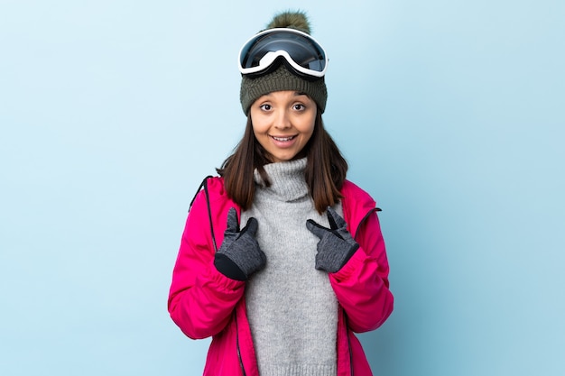 Gemischte Rennskifahrerin mit Snowboardbrille über isoliertem Blau mit überraschendem Gesichtsausdruck.