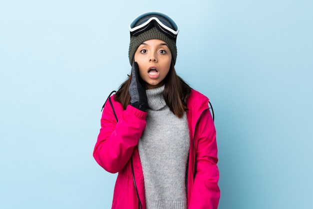 Gemischte Rennskifahrerin mit Snowboardbrille über isoliertem Blau mit Überraschung und schockiertem Gesichtsausdruck.