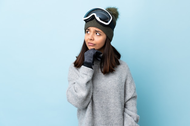 Gemischte Rennskifahrerin mit Snowboardbrille über isoliertem blauem Raum, der Zweifel und mit verwirrendem Gesichtsausdruck hat