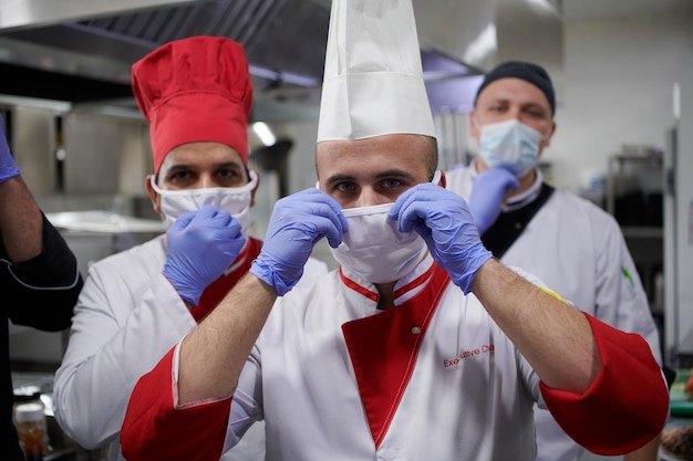 Gemischtes Rennteam Porträt von Gruppenköchen, die in der Küche des Restaurants zusammenstehen und eine medizinische Schutzmaske und Handschuhe im neuen normalen Konzept des Coronavirus tragen