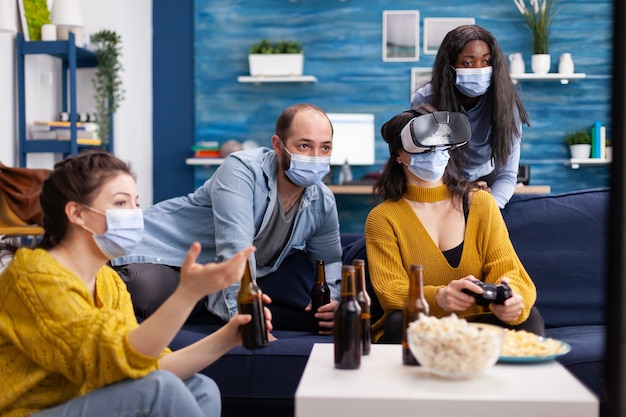 Gemischtrassige Freunde im Wohnzimmer, die einen Videospielwettbewerb haben, halten die soziale Distanzierung mit Gesichtsmaske, um eine Krankheit mit Covid zu verhindern. Virtuelle Realität erleben. Konzeptionelles Bild.