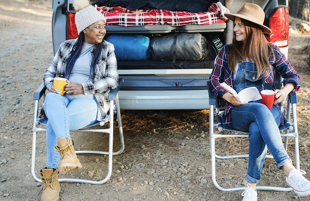 Gemischtrassige Freundinnen, die Spaß beim Camping mit Wohnmobil haben, während sie lesen und Kaffee im Freien trinken Fokus auf Gesichter
