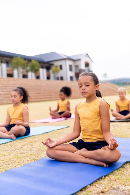 Gemischtrassige Grundschülerinnen meditieren, während sie auf dem Schulgelände auf einer Gymnastikmatte sitzen
