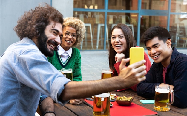 Gemischtrassige Gruppe von Freunden in einer Bar, die Bier trinken, schauen sich gemeinsam das Handy an Videoanruf