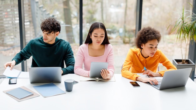 Gemischtrassige Studenten sitzen im Universitätspublikum und nutzen verschiedene Geräte, um drinnen für Prüfungen zu lernen