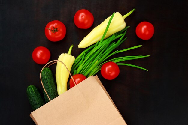 Gemüse fiel aus einer Papiertüte auf dunklem Hintergrund Draufsicht Das Konzept der Lebensmittel in Säcken Vegetarische gesunde Lebensmittel vom Markt Frühlingszwiebeln Tomaten und gelbe Paprika