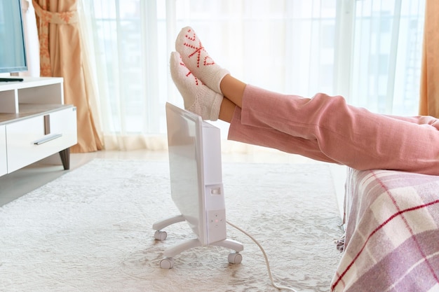 Gemütliche Frau mit bequemen, weichen Stricksocken bekommt wärmende Füße mit moderner weißer tragbarer elektrischer Heizung zu Hause