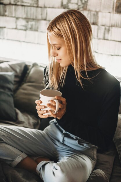 Gemütliche junge Frau, die heißen Kaffee trinkt, während sie zu Hause im Bett sitzt.