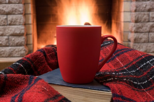 Gemütliche Szene vor Kamin mit rotem Becher mit Tee, Buch, Wollschal.