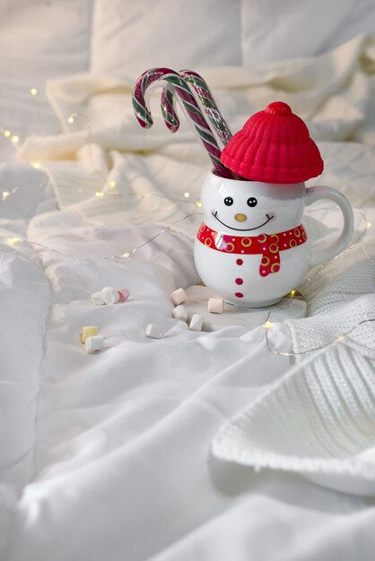 Gemütliche weiche Decke mit einer Tasse in Form eines Schneemanns mit Weihnachtssüßigkeiten auf dem Bett