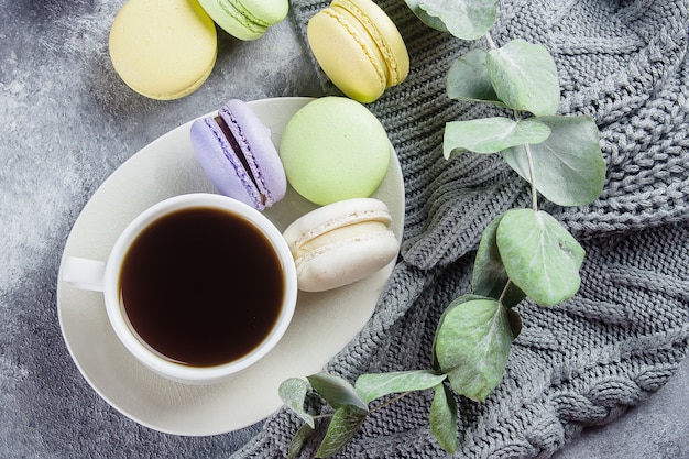 Gemütliches Morgen-Konzept. Köstliche bunte Pastell-Macarons mit Sahne und Kaffee, warme graue Strickjacke