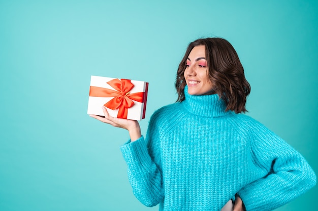 Gemütliches Porträt einer jungen Frau in einem gestrickten blauen Pullover und leuchtend rosa Make-up mit einer Geschenkbox