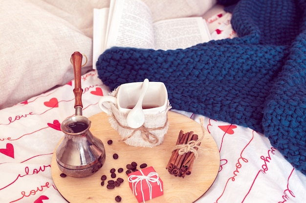 Gemütliches Stillleben mit Frühstück im Bett zum Valentinstag. Kaffee mit kleinem Geschenk und Plaid mit Buch auf einem Hintergrund in der Isolationszeit