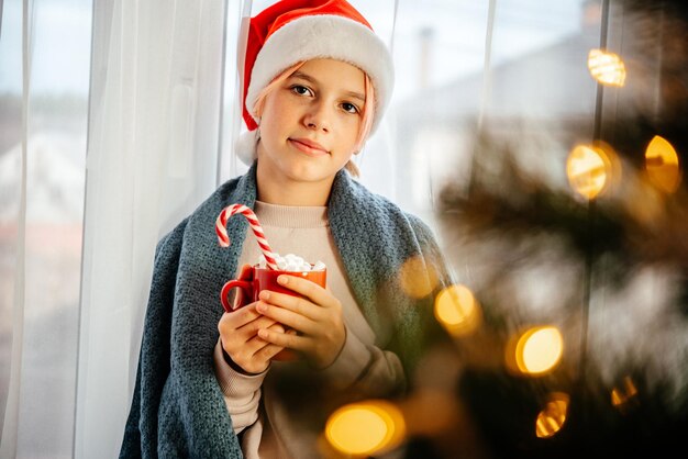Gemütliches Weihnachten zu Hause festlicher Hintergrund mit einem Mädchen in Weihnachtsmütze und einer Tasse heißer Schokolade