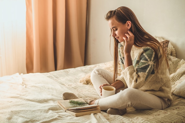 Gemütliches zu Hause. Schöne Frau liest ein Buch auf dem Bett. Guten Morgen mit Tee und Buch. Hübsche junge Frau, die sich entspannt. Das Konzept des Lesens