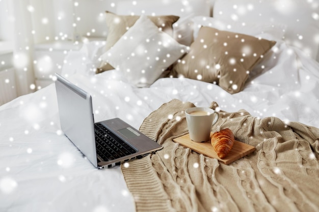 gemütliches Zuhause, Interieur und Winterkonzept - gemütliches Schlafzimmer mit Laptop-Computer, Kaffeetasse und Croissant im Bett zu Hause über Schnee