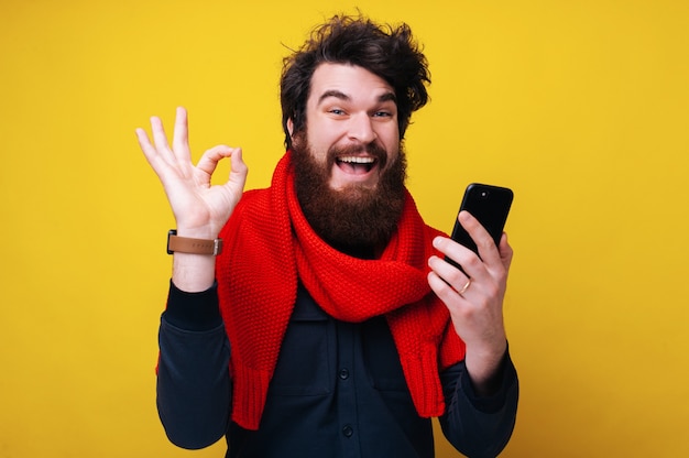 Genehmigt! Foto eines fröhlichen bärtigen Mannes, der ein Mobiltelefon hält und ein OK-Zeichen zeigt, der auf gelbem, isoliertem Hintergrund steht