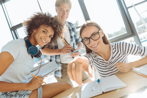 Generación inteligente, jóvenes estudiantes alegres que le señalan mientras están sentados en la mesa y estudian juntos